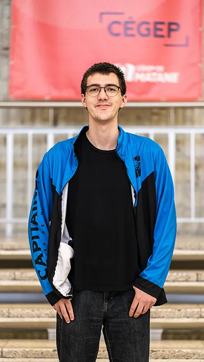 Un athlète de l'équipe de sport électronique pose sur les marches de l'escalier du hall d'entrée du Cégep de Matane pour sa photo d'équipe, vêtu de la veste bleue et noire des Capitaines.