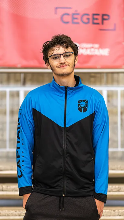 Un athlète de l'équipe de sport électronique pose sur les marches de l'escalier du hall d'entrée du Cégep de Matane pour sa photo d'équipe, vêtu de la veste bleue et noire des Capitaines.