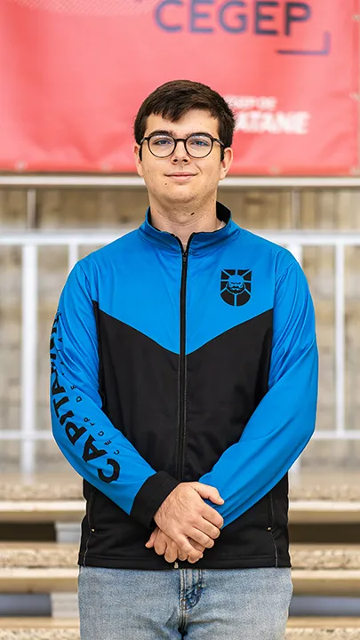 Un athlète de l'équipe de sport électronique pose sur les marches de l'escalier du hall d'entrée du Cégep de Matane pour sa photo d'équipe, vêtu de la veste bleue et noire des Capitaines.