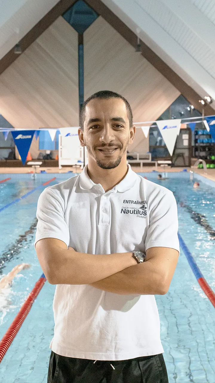 Devant le bassin d'une piscine, un entraîneur de natation en tenue de sport prend la pose, dans le cadre de la prise des photos officielles de l'équipe de natation des Capitaines.