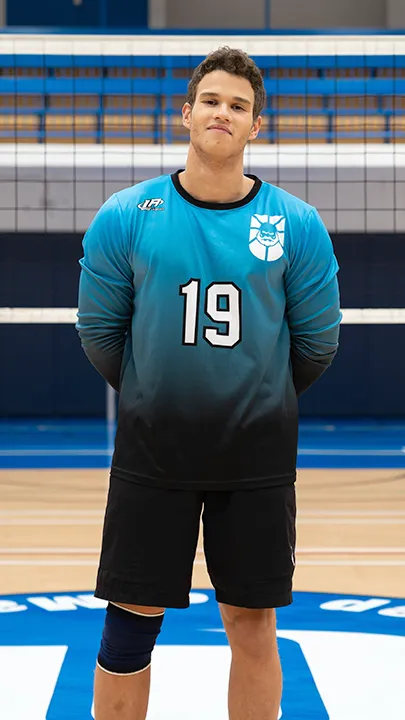 Dans un gymnase, devant un filet de volleyball installé sur un terrain omnisport, un athlète en tenue officielle des Capitaines pose lors de la prise des photos officielles de l'équipe.