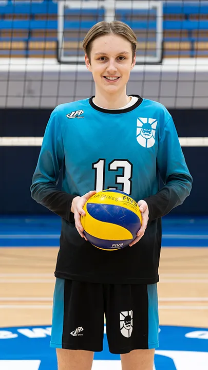 Dans un gymnase, devant un filet de volleyball installé sur un terrain omnisport, un athlète en tenue officielle des Capitaines pose avec un ballon de volleyball lors de la prise des photos officielles de l'équipe.