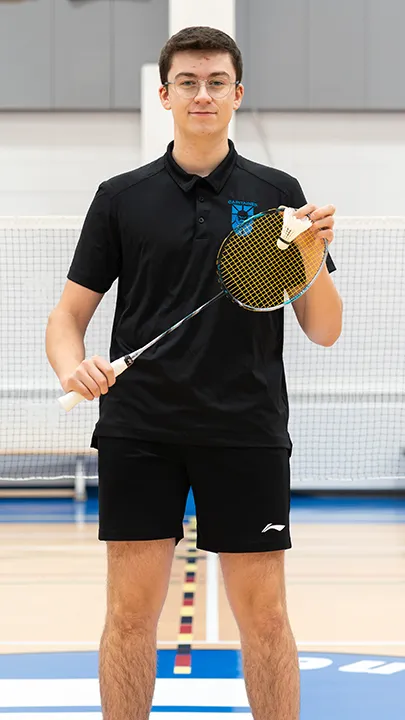 Sur un terrain de badminton, dans un gymnase, un athlète de l'équipe des Capitaines, en tenue officielle, une raquette de badminton à la main, prend la pose dans le cadre des photos de l'équipe.