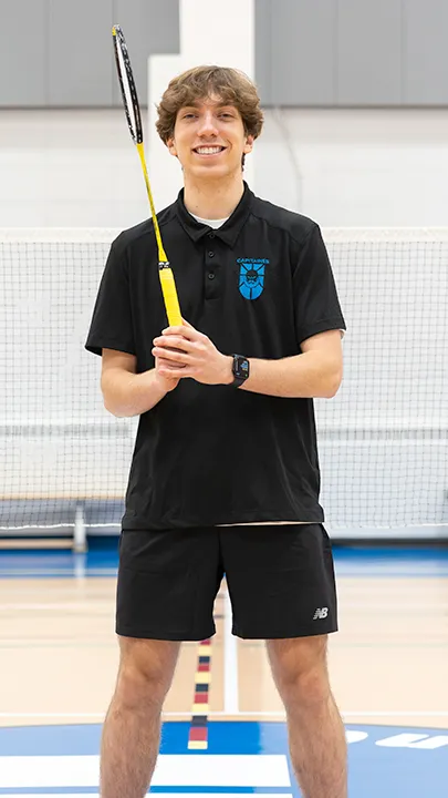 Sur un terrain de badminton, dans un gymnase, un athlète de l'équipe des Capitaines, en tenue officielle, une raquette de badminton à la main, prend la pose dans le cadre des photos de l'équipe.