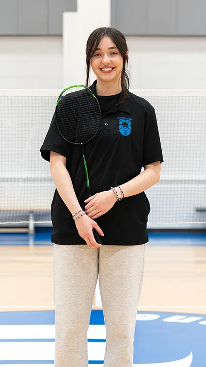 Sur un terrain de badminton, dans un gymnase, une athlète de l'équipe des Capitaines, en tenue officielle, une raquette de badminton à la main, prend la pose dans le cadre des photos de l'équipe.