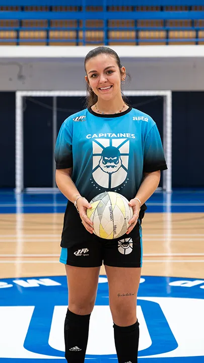 Dans un gymnase, une athlète étudiante du cégep pose seule, en tenue bleue et noire des Capitaines, un ballon à la main, dans le cadre des photos d'équipe de futsal féminin pour l'année 2024-2025.