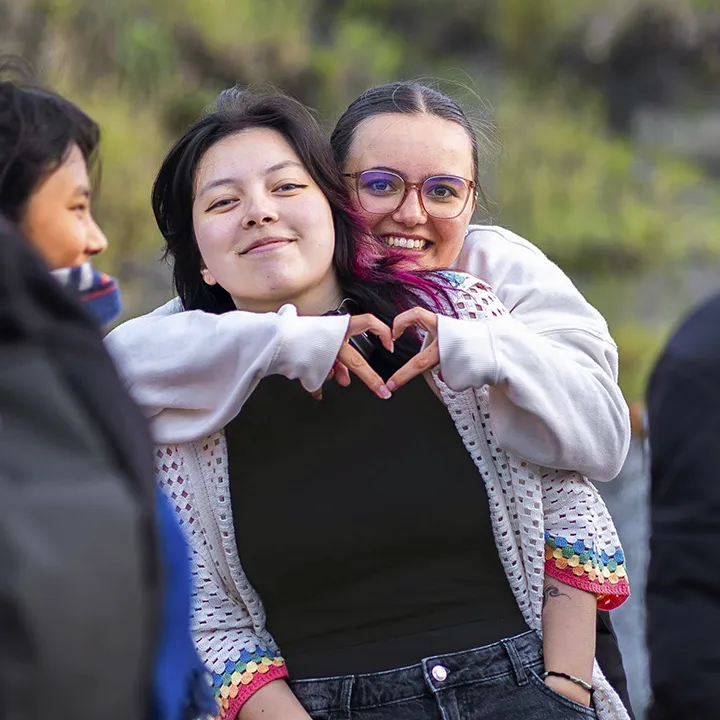 Deux amies, dans une foule d'étudiantes et d'étudiants qu'on devine, profitent d'une activité sociale en plein air, le sourire aux lèvres. L'une d'entre elles, les bras autour de son amie, fait la forme d'un cœur avec ses doigts.