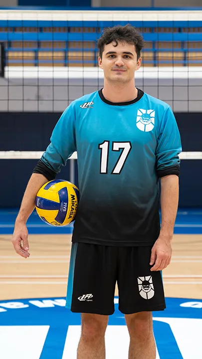 Dans un gymnase, devant un filet de volleyball installé sur un terrain omnisport, un athlète en tenue officielle des Capitaines pose avec un ballon de volleyball lors de la prise des photos officielles de l'équipe.