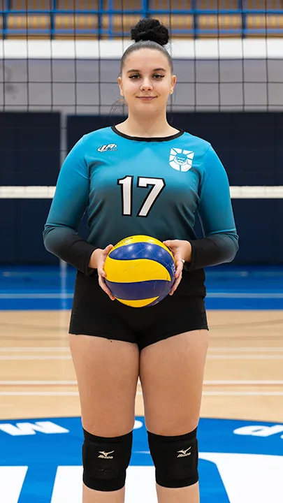 Sur le terrain omnisport d'un gymnase, devant un filet de volleyball, une étudiante vêtue de l'uniforme des Capitaines prend la pose, un ballon de volleyball entre les mains, dans le cadre des photos officielles de l'équipe féminine de volleyball du cégep.