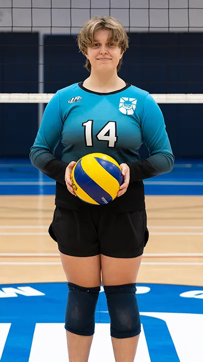 Sur le terrain omnisport d'un gymnase, devant un filet de volleyball, une étudiante vêtue de l'uniforme des Capitaines prend la pose, un ballon de volleyball entre les mains, dans le cadre des photos officielles de l'équipe féminine de volleyball du cégep.