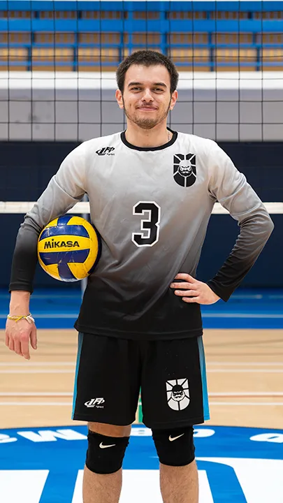 Dans un gymnase, devant un filet de volleyball installé sur un terrain omnisport, un athlète en tenue officielle des Capitaines pose avec un ballon de volleyball lors de la prise des photos officielles de l'équipe.