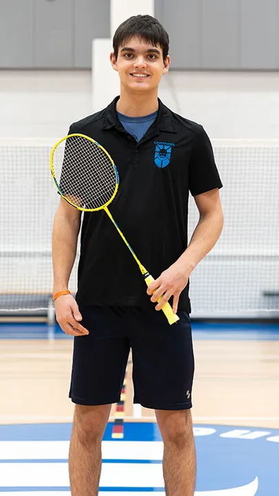 Sur un terrain de badminton, dans un gymnase, un athlète de l'équipe des Capitaines, en tenue officielle, une raquette de badminton à la main, prend la pose dans le cadre des photos de l'équipe.