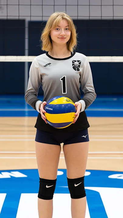 Sur le terrain omnisport d'un gymnase, devant un filet de volleyball, une étudiante vêtue de l'uniforme des Capitaines prend la pose, un ballon de volleyball entre les mains, dans le cadre des photos officielles de l'équipe féminine de volleyball du cégep.