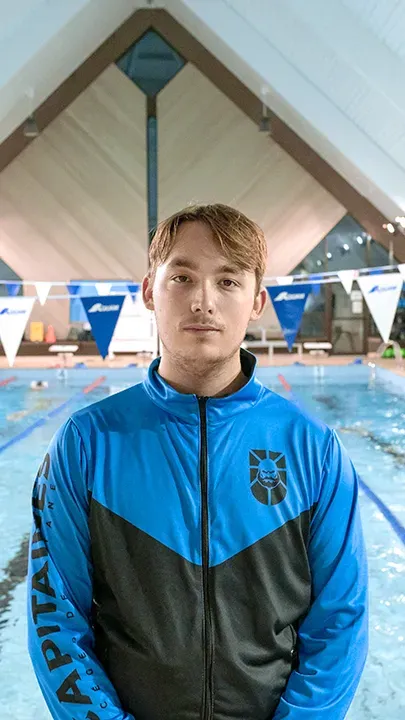 Un athlète des Capitaines, dans une piscine, sur le bord d'un bassin de natation, pose avec la tenue bleue et noire des Capitaines dans le cadre de la photo officielle de l'équipe collégiale de natation.