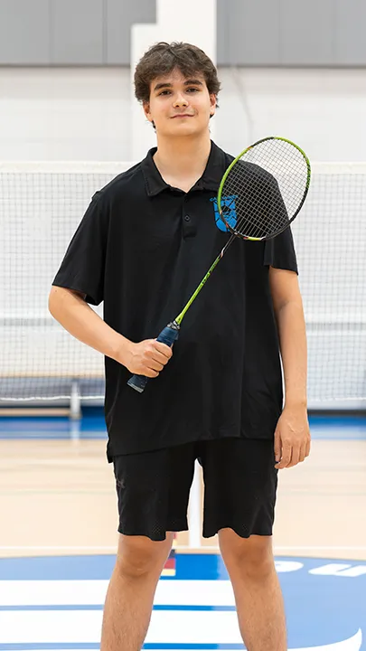 Sur un terrain de badminton, dans un gymnase, un athlète de l'équipe des Capitaines, en tenue officielle, une raquette de badminton à la main, prend la pose dans le cadre des photos de l'équipe.