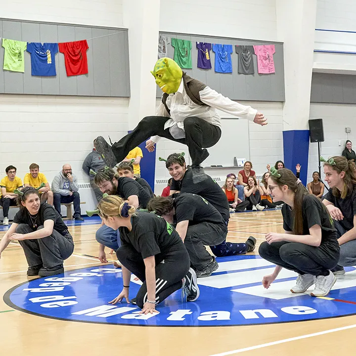 Dans un gymnase, des étudiantes et étudiants effectuent une chorégraphie devant un public assis sur le banc de touche, dans le cadre des Olympiades de la vie active organisées par la vie étudiante. La mascotte de l'équipe, déguisée en Shrek, sautent par-dessus ses coéquipiers sous les regards impressionnés du public.