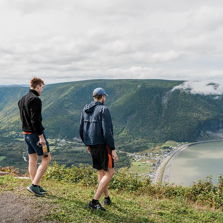 Deux étudiants en tenue de sport dominent un panorama à couper le souffle au sommet du mont Saint-Pierre. Depuis le haut de cette montagne à gravir en Gaspésie, on aperçoit le village en contrebas, le fleuve et quelques nuages accrochés aux sommets juste en face.