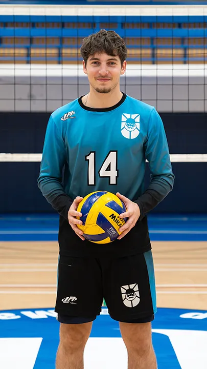 Dans un gymnase, devant un filet de volleyball installé sur un terrain omnisport, un athlète en tenue officielle des Capitaines pose avec un ballon de volleyball lors de la prise des photos officielles de l'équipe.