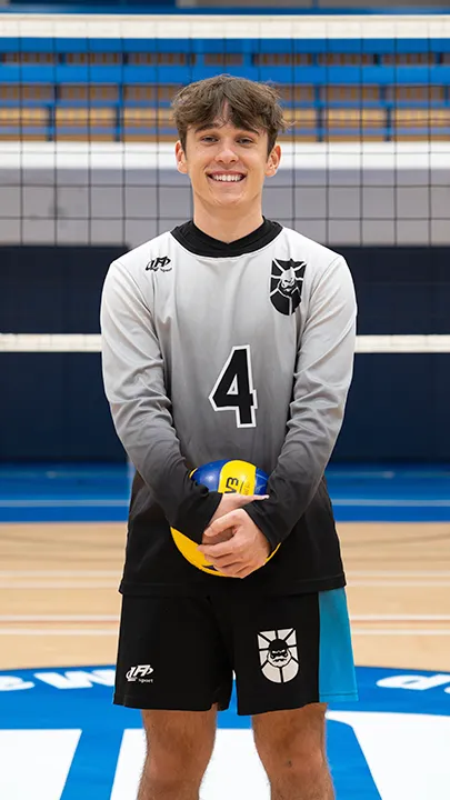 Dans un gymnase, devant un filet de volleyball installé sur un terrain omnisport, un athlète en tenue officielle des Capitaines pose avec un ballon de volleyball lors de la prise des photos officielles de l'équipe.