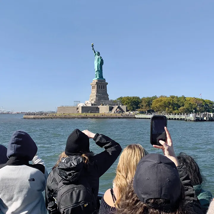 Sur le pont d'un bateau, des étudiantes et étudiants brandissent leurs appareils photos et téléphones portables alors que la statue de la Liberté, à New York, se dégage devant eux sur un ciel bleu.