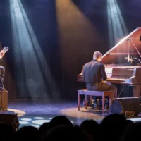 Trois étudiants performent sur scène lors de Cégeps en spectacle : un étudiant joue de la guitare acoustique, un autre est au piano à queue et une étudiante chante, un micro à la main.