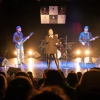 Un groupe de musique avec un batteur, deux guitaristes et une chanteuse assure un spectacle sur la scène de l'auditorium du cégep.