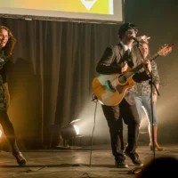 Deux enseignantes sont en train de danser le twist pendant qu'un guitariste déguisé en Beatles, une perruque noire sur la tête, des lunettes rondes devant les yeux, en veston et cravate, est en train de jouer de la guitare.