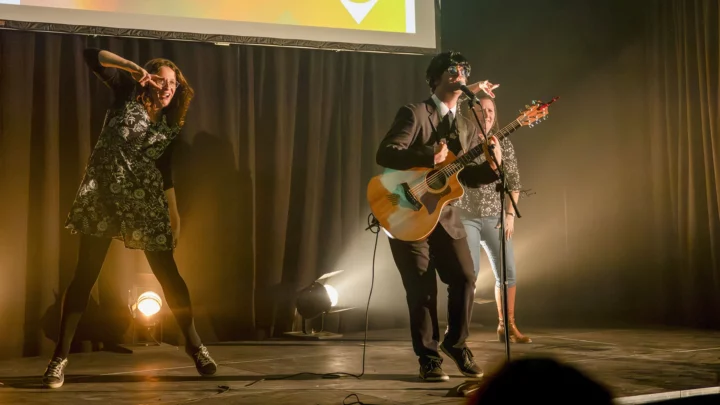 Deux enseignantes sont en train de danser le twist pendant qu'un guitariste déguisé en Beatles, une perruque noire sur la tête, des lunettes rondes devant les yeux, en veston et cravate, est en train de jouer de la guitare.