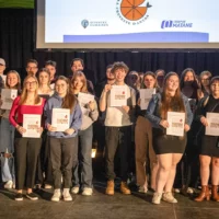 Sur une scène, plusieurs étudiantes et étudiants, ainsi que leur enseignante, prennent la pose, un exemplaire de La Crevette d'antan à la main, pour immortaliser le dévoilement de leur journal étudiant.