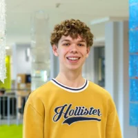 Un étudiant se tient dans le hall d'entrée du cégep pour une prise de photo officielle soulignant son rôle d'ambassadeur du programme Sciences humaines. Il porte un chandail jaune et affiche un grand sourire.