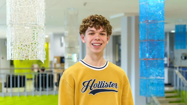 Un étudiant se tient dans le hall d'entrée du cégep pour une prise de photo officielle soulignant son rôle d'ambassadeur du programme Sciences humaines. Il porte un chandail jaune et affiche un grand sourire.